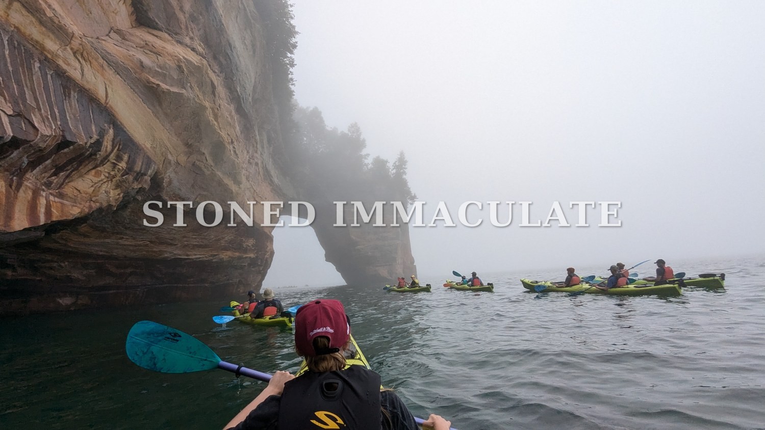 A line of green kayaks follows the stained sandstone cliffs toward an arch in thick fog on Lake Superior