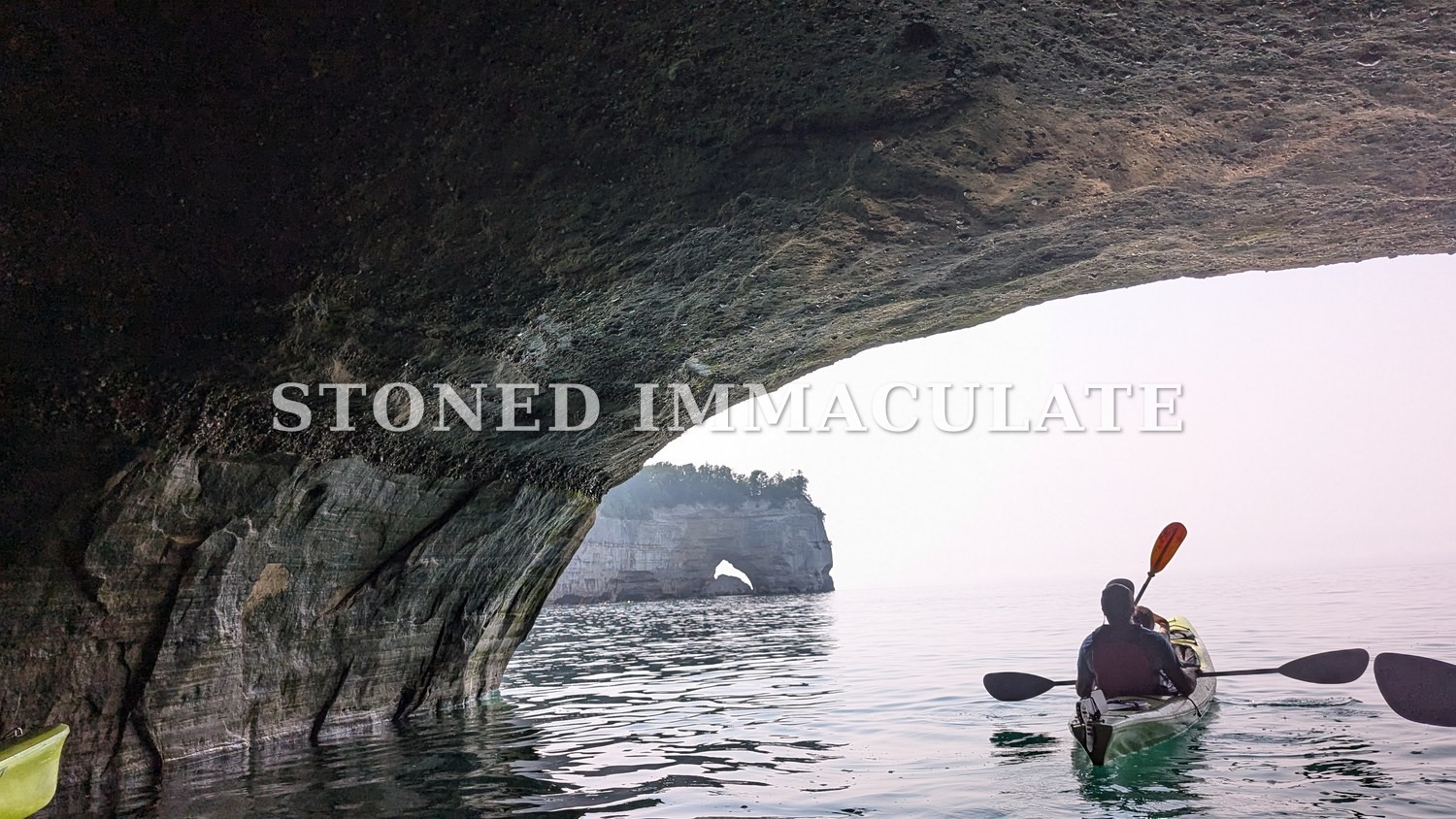 A kayaker paddles out of the dark of a sea cave toward a distant rock arch on Lake Superior